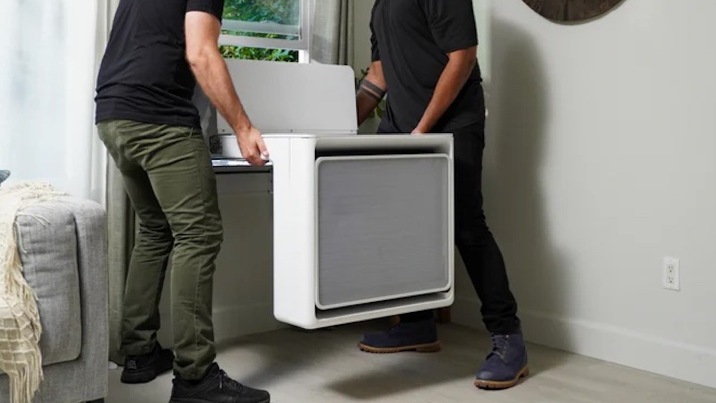 Two people are installing a modern heat pump unit near a window in a well-lit room.