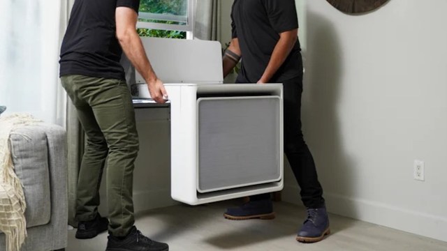 Two people are installing a modern heat pump unit near a window in a well-lit room.