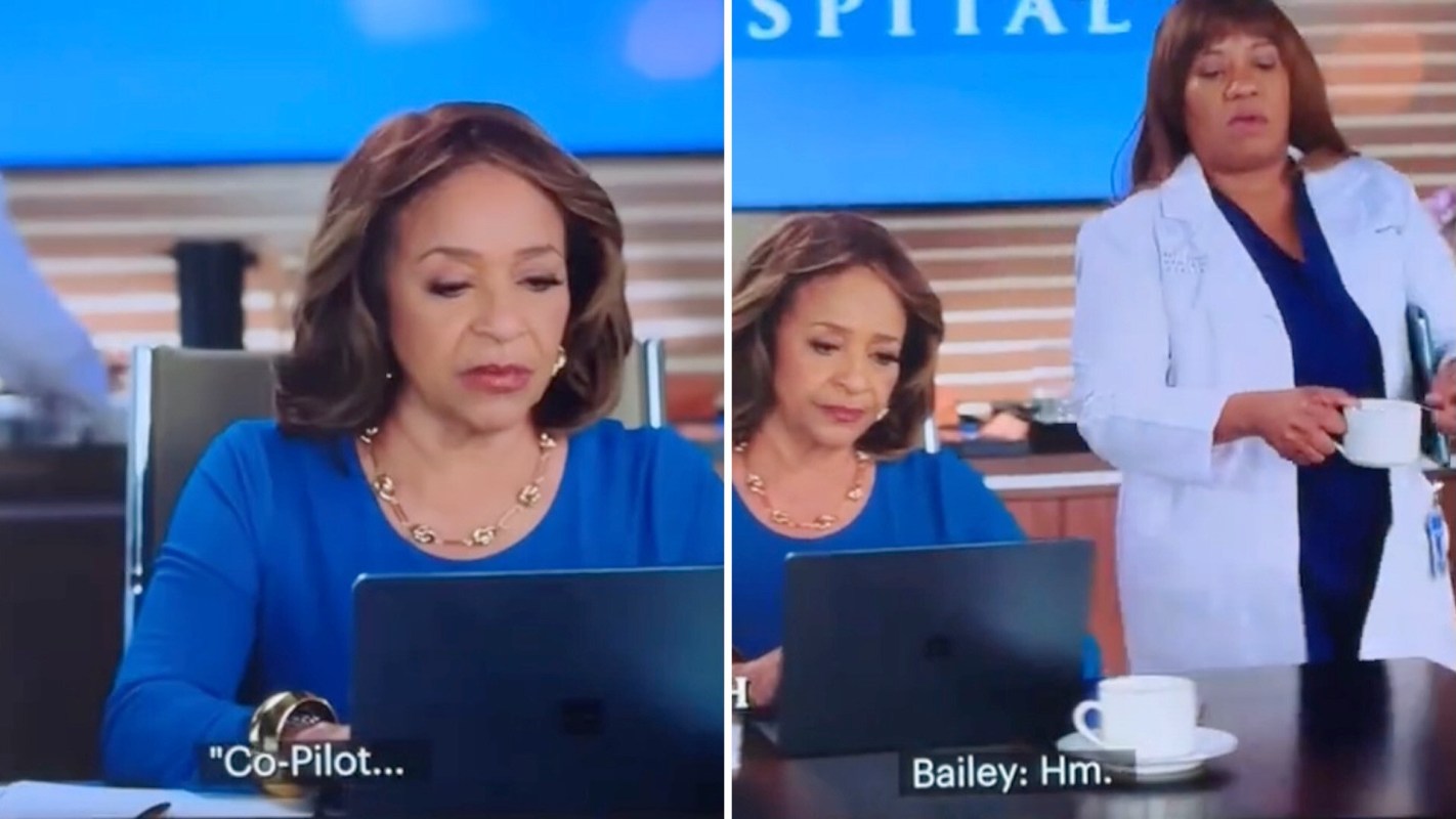 A woman in a blue shirt looks thoughtfully at her laptop while another woman in a white coat holds a cup nearby.