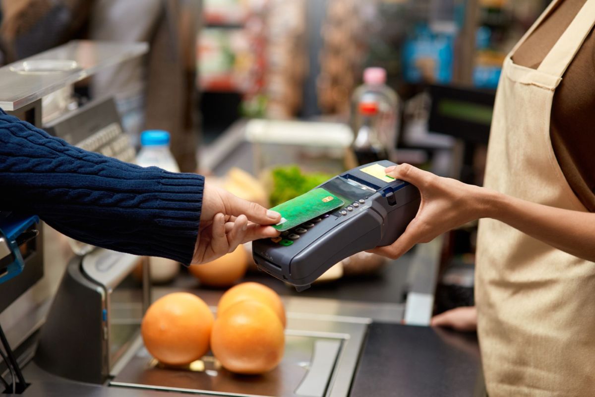 A customer makes a payment at a grocery store checkout using a card reader, with oranges in the foreground.