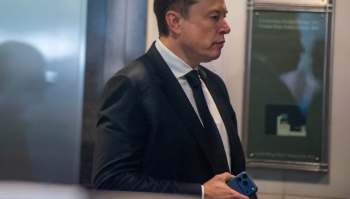 An unsmiling Elon Musk looks to the right, holding a smartphone.