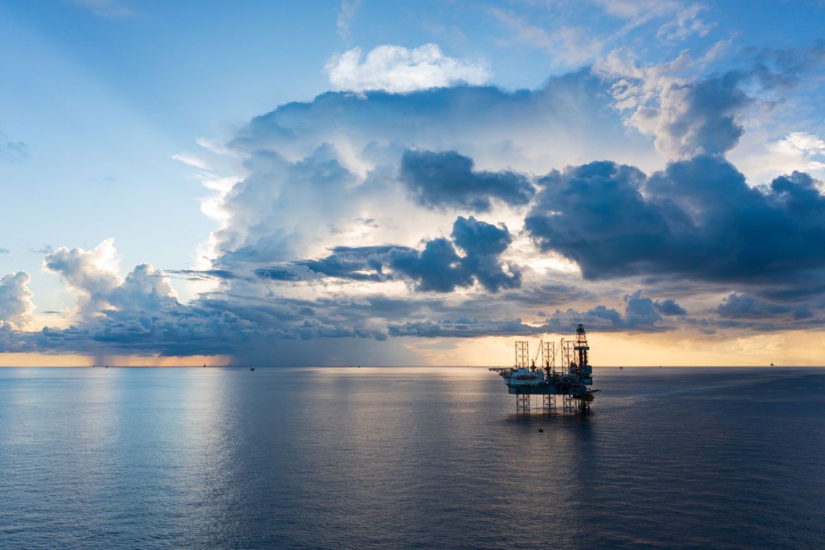 An offshore oil rig stands in calm waters under a dramatic sky with fluffy clouds and a sunset backdrop.