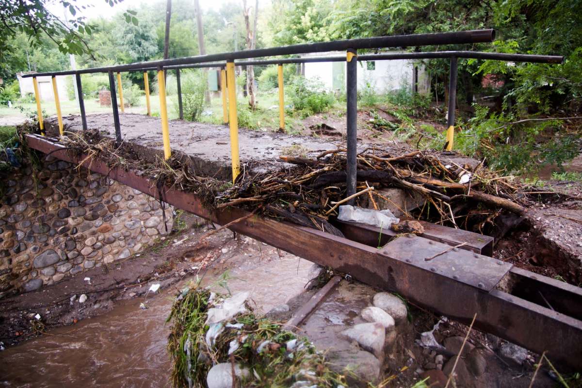 A damaged metal bridge covered with debris spans a shallow, muddy waterway in a wooded area.