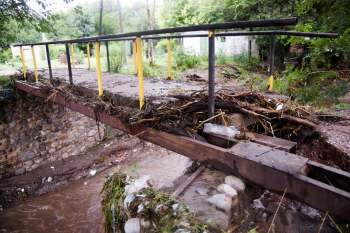 A damaged metal bridge covered with debris spans a shallow, muddy waterway in a wooded area.