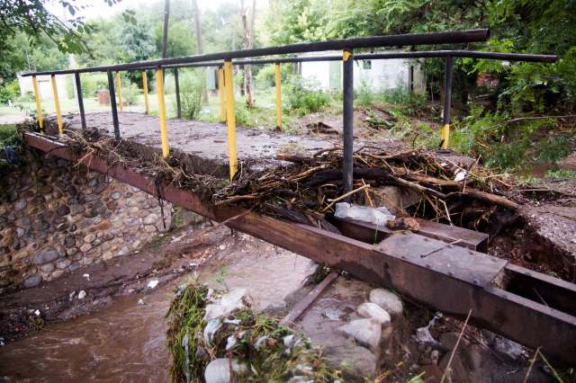 A damaged metal bridge covered with debris spans a shallow, muddy waterway in a wooded area.