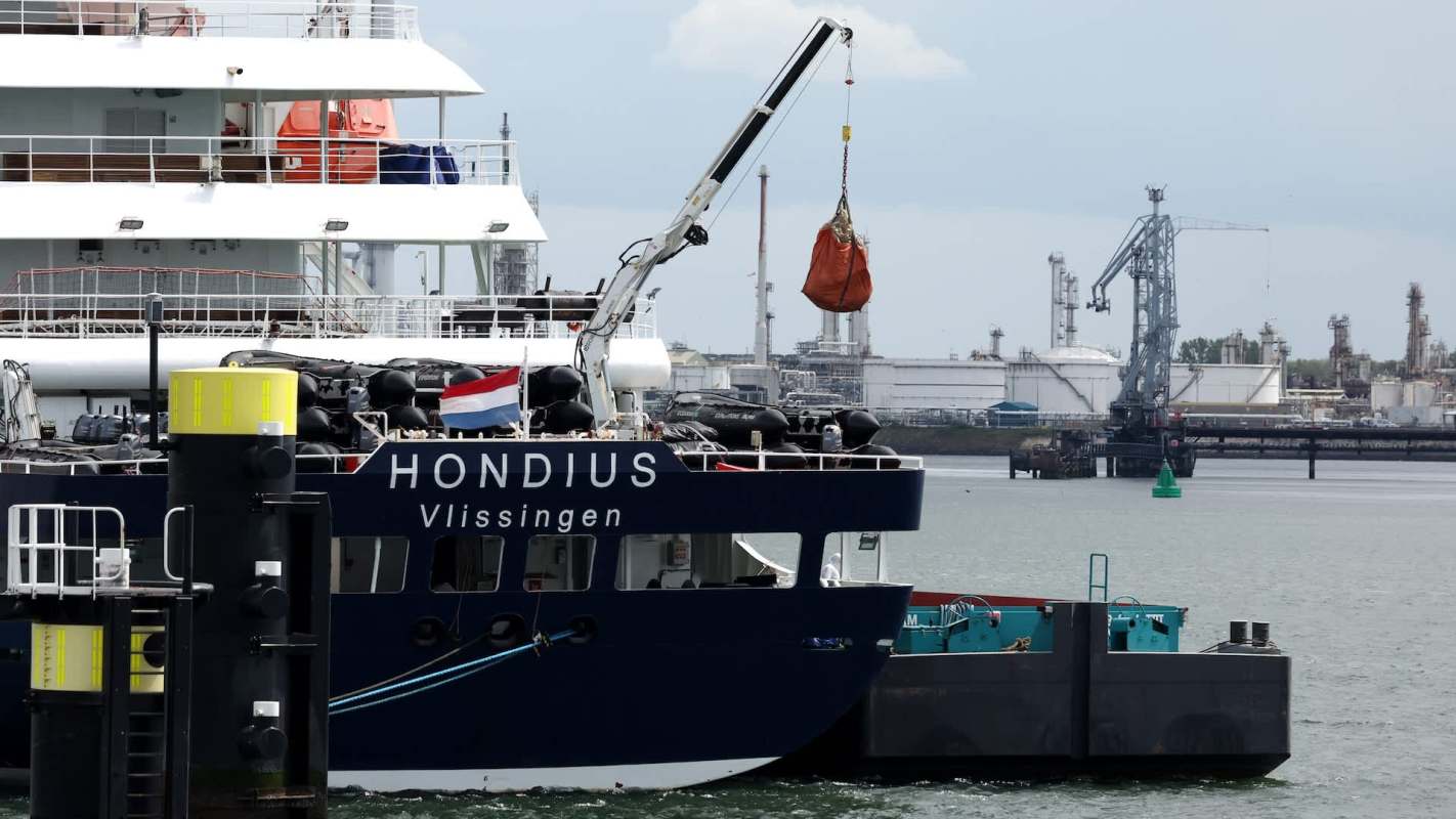 A cargo ship named Hondius is docked, with a crane lifting a load at a busy industrial port.