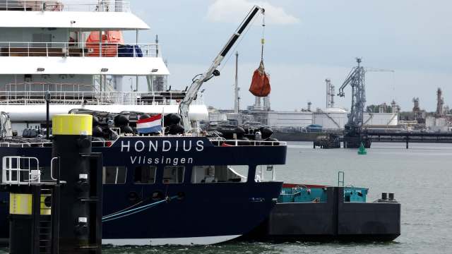 A cargo ship named Hondius is docked, with a crane lifting a load at a busy industrial port.