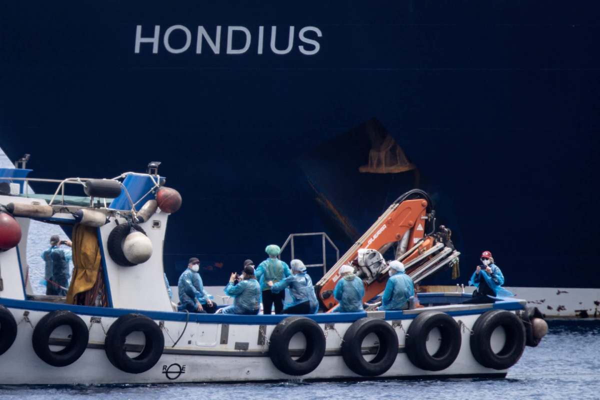 A small boat carries several people in blue jackets near a large ship named "Hondius."