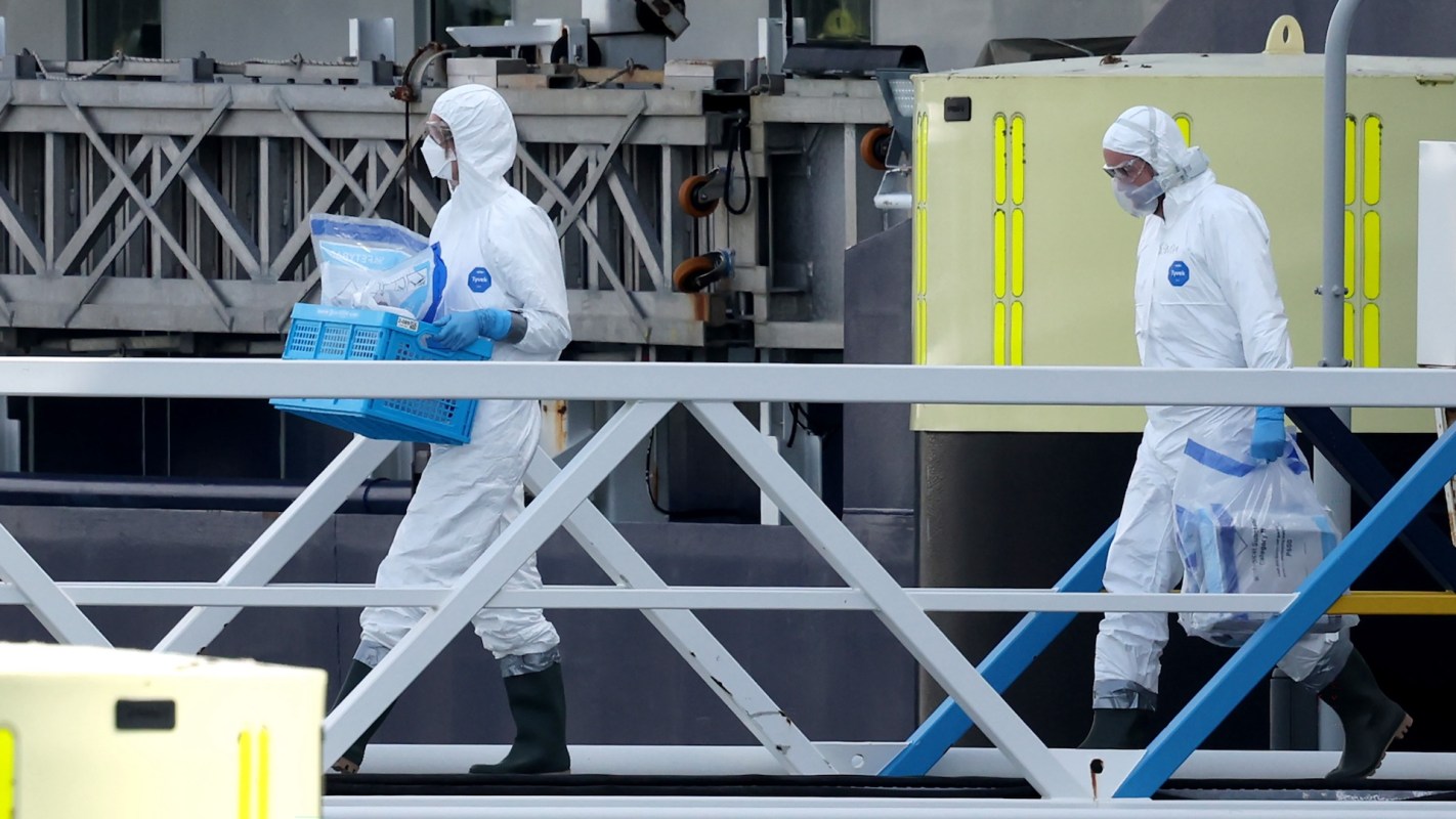 Two people in white protective suits carrying equipment walk along a dock near a large industrial structure.
