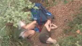 A person lies on the ground beside a backpack in a wooded area, while another person is nearby.