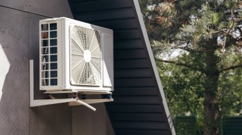 A white heat pump unit mounted on a wall with greenery in the background.
