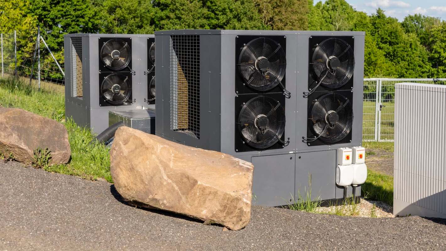 A large industrial cooling unit with multiple fans sits on a gravel surface, surrounded by grass and boulders.