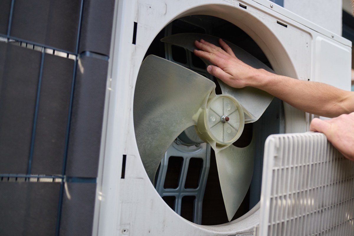 A person inspecting the fan inside a heat pump unit with one hand.