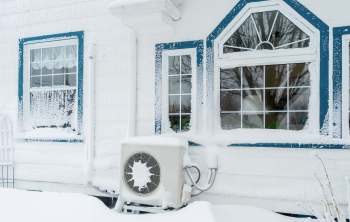 A house exterior covered in heavy snow, featuring frosted windows and a snow-covered outdoor heat pump.