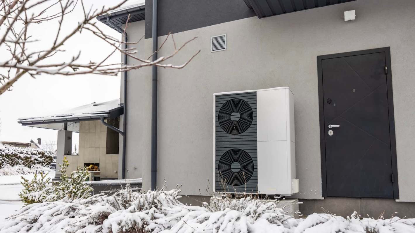 An outdoor heat pump unit beside a dark door, set against a snowy landscape.