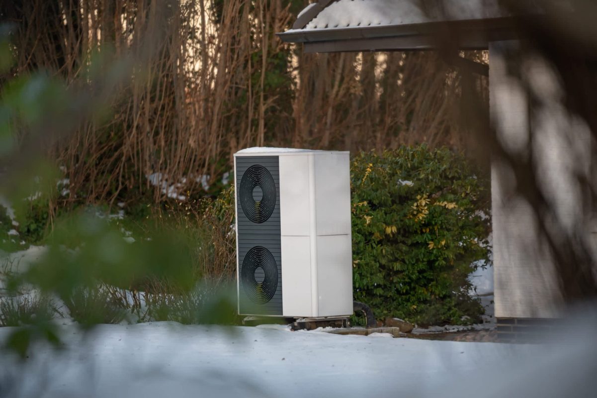 A heat pump unit stands outside in a snowy landscape, surrounded by greenery and a building.