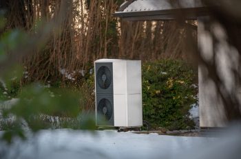A heat pump unit stands outside in a snowy landscape, surrounded by greenery and a building.