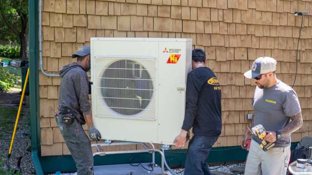 Three technicians are installing a Mitsubishi heat pump by a wooden shingle wall.