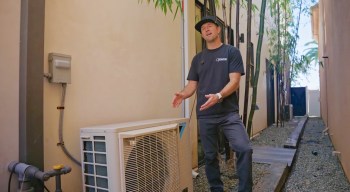 A man standing beside an outdoor air conditioning unit in a narrow alleyway surrounded by bamboo.