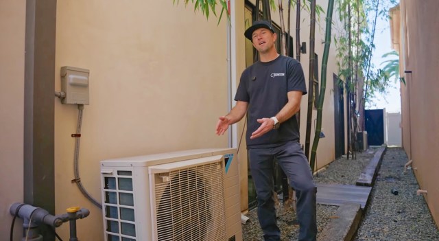 A man standing beside an outdoor air conditioning unit in a narrow alleyway surrounded by bamboo.