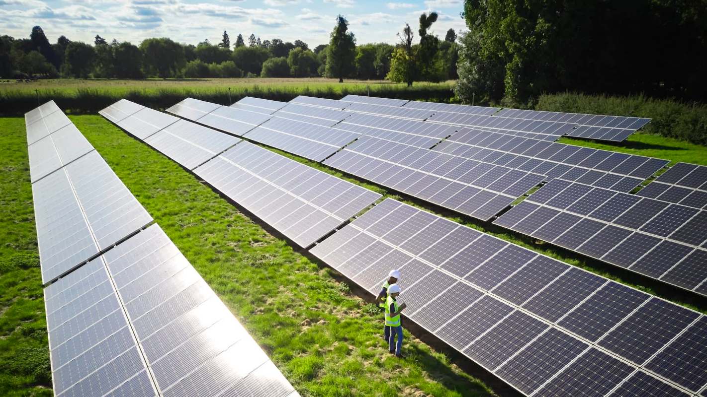 Two workers in safety gear inspect solar panels arranged in rows on a grassy field.