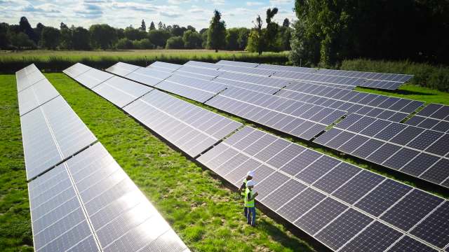 Two workers in safety gear inspect solar panels arranged in rows on a grassy field.