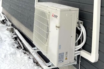 A wall-mounted heat pump outside a house surrounded by snow.