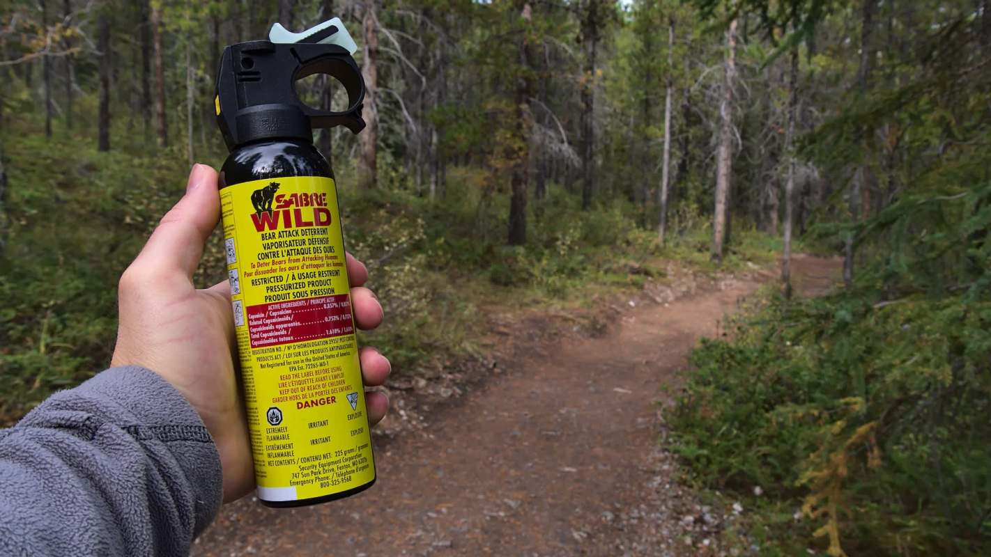 A hand holding a bear spray canister on a forested hiking trail.