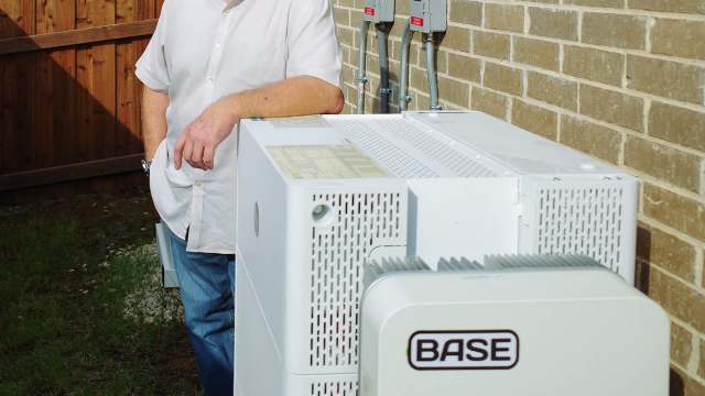 A man stands next to a large white utility box against a brick wall and wooden fence.