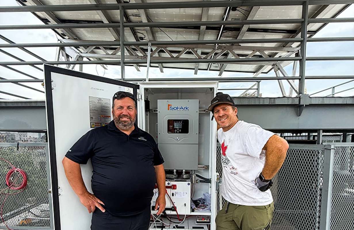 Two men stand smiling beside a solar power unit with a gray cabinet and solar panels above them.