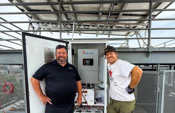Two men stand smiling beside a solar power unit with a gray cabinet and solar panels above them.