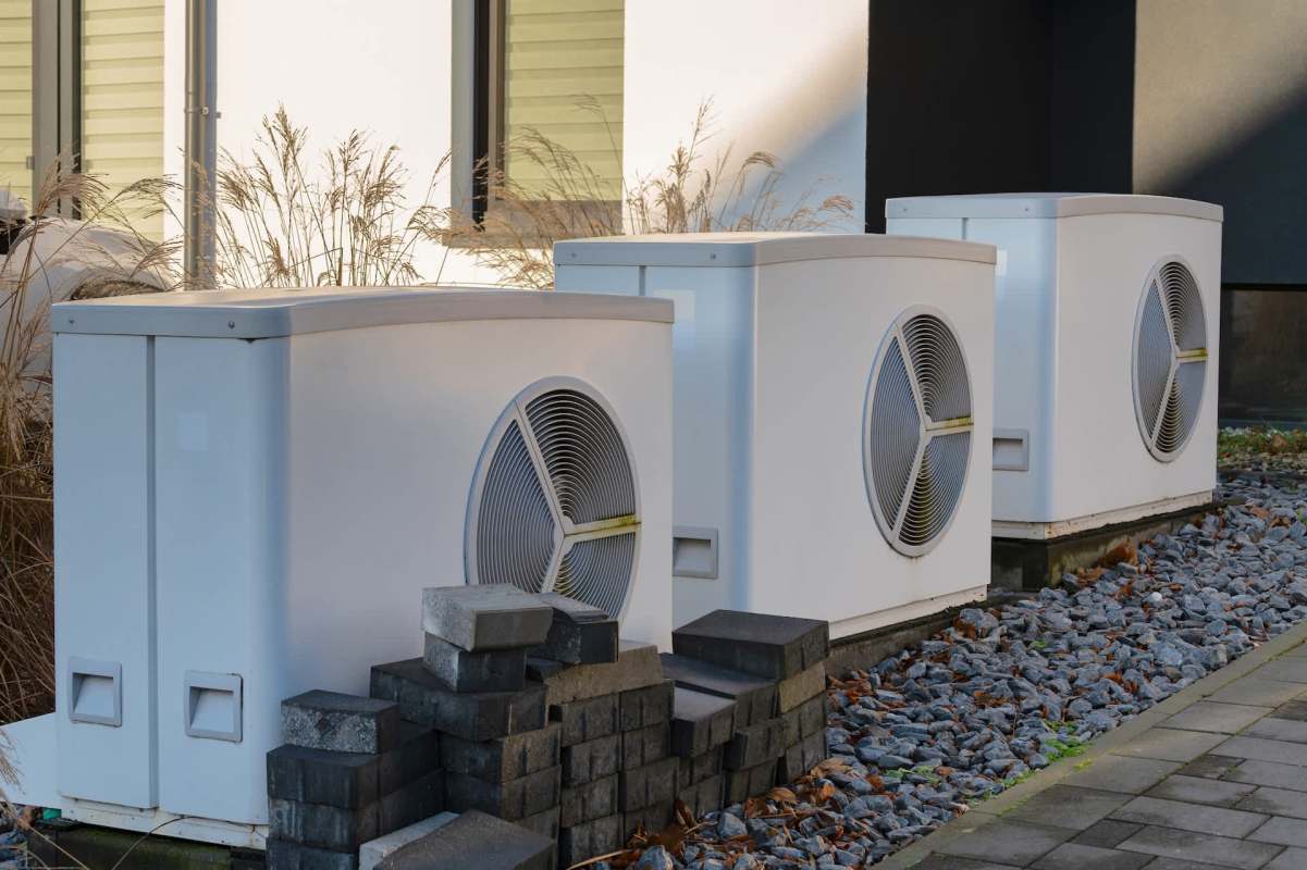 Three white heat pump units sit on a gravel bed beside a modern building with grass and fallen leaves.