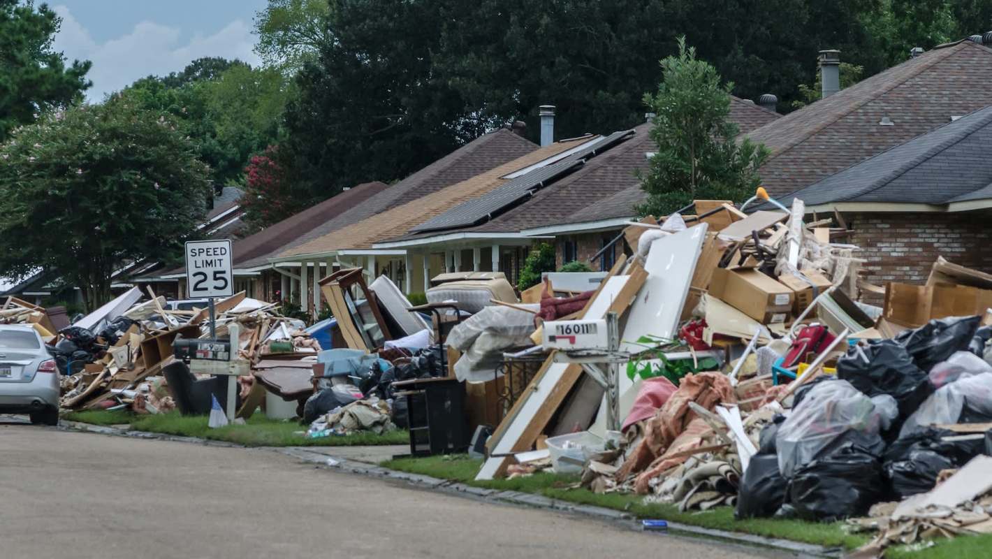 Street lined with homes, piled with debris and discarded furniture after a flood, with a speed limit sign nearby.