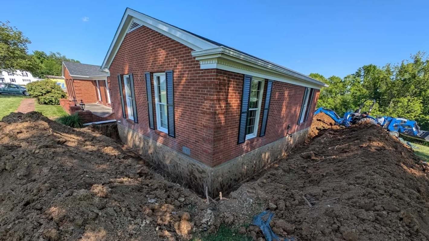A brick house with a deep excavation around its foundation and heavy machinery in the background.