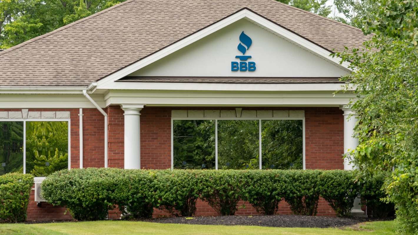 A brick building featuring a "BBB" sign for the Better Business Bureau, surrounded by lush green bushes and trees.