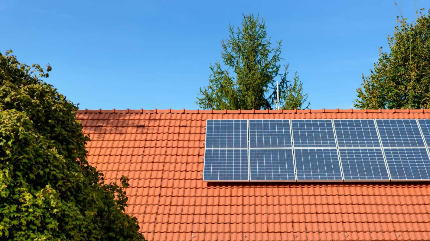 A red-tiled roof with solar panels installed, surrounded by greenery under a clear blue sky.