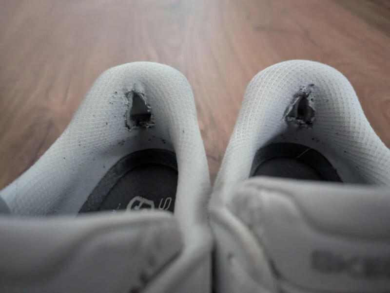 A pair of white sneakers with holes in the inner heel fabric. 
