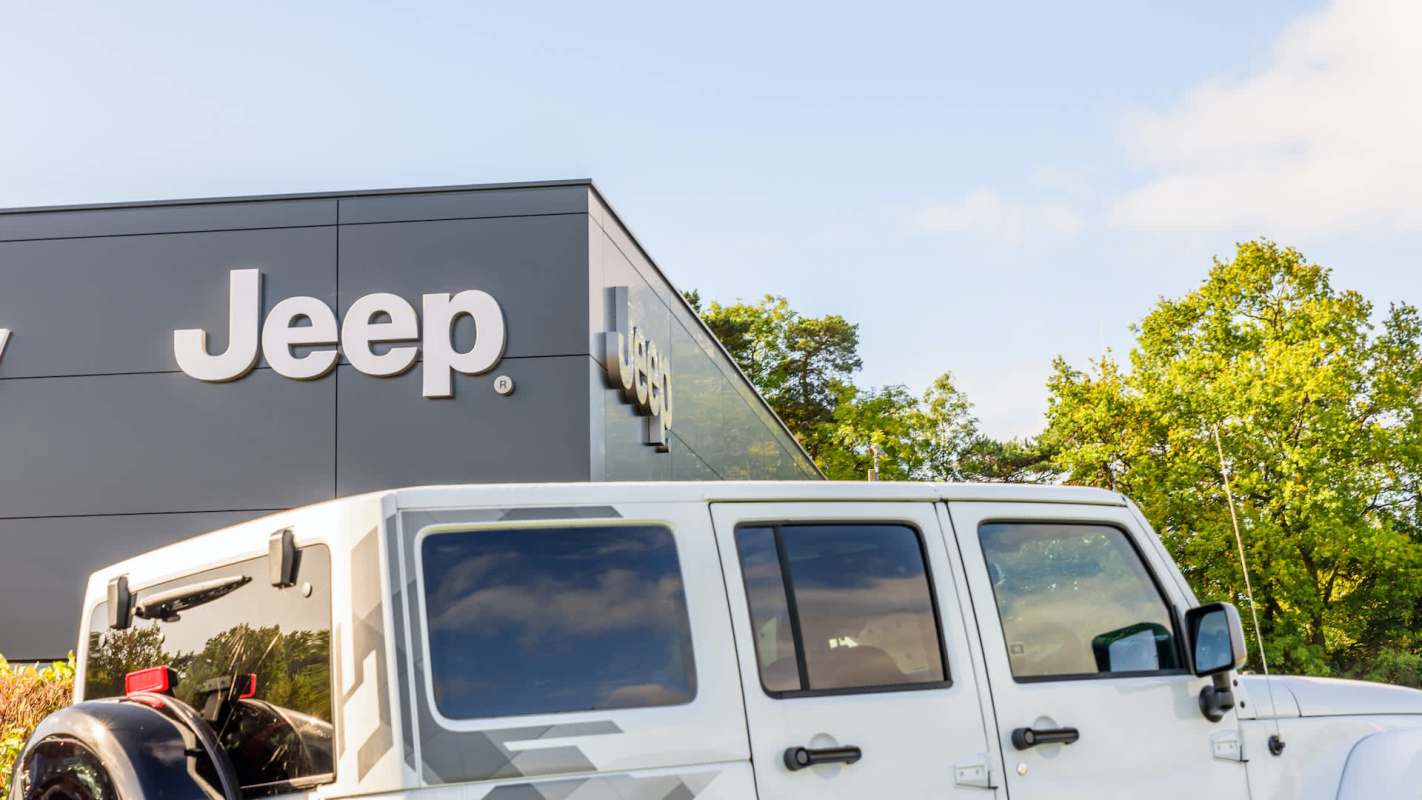 A white Jeep parked in front of a modern Jeep dealership building with a clear blue sky.