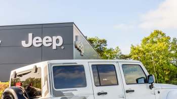 A white Jeep parked in front of a modern Jeep dealership building with a clear blue sky.