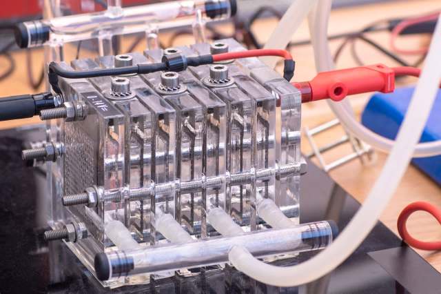 A close-up of a clear, multi-chambered electrolysis cell with connected tubing and wires.