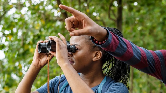 A woman uses binoculars outdoors while a person points out something in the distance.