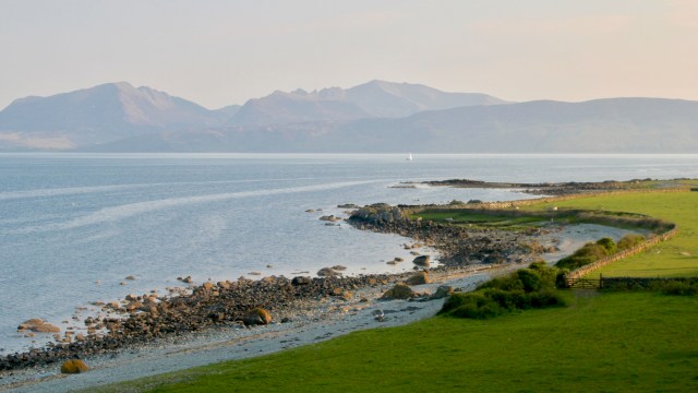 A coastal scene showcasing rocky shores, green fields, and distant mountains under a clear sky.