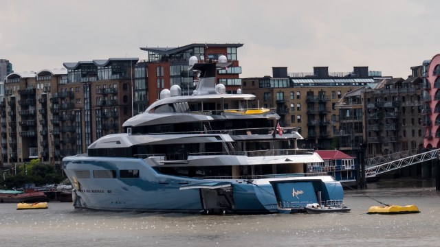 A large luxury yacht named "Aviva" with a British flag docked by modern buildings along a river.