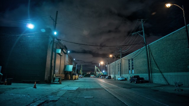 An empty industrial alleyway at night, illuminated by streetlights and lined with parked vehicles and utility poles.
