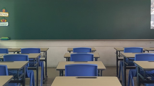 A classroom with blue chairs and tables facing a green chalkboard, with minimal decorations on the wall.