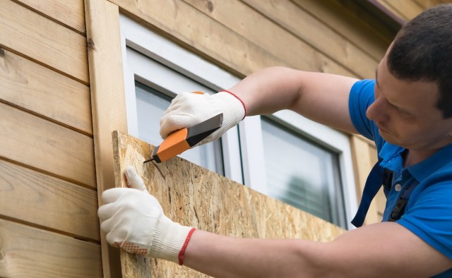 A person in gloves measures and cuts a plywood board to fit over a window.