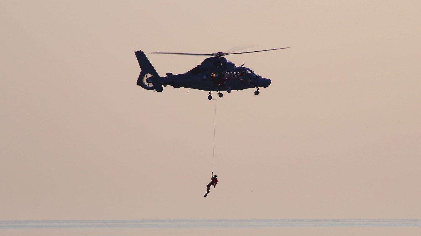 A helicopter hovers above water while a rescue diver is lowered on a rope.