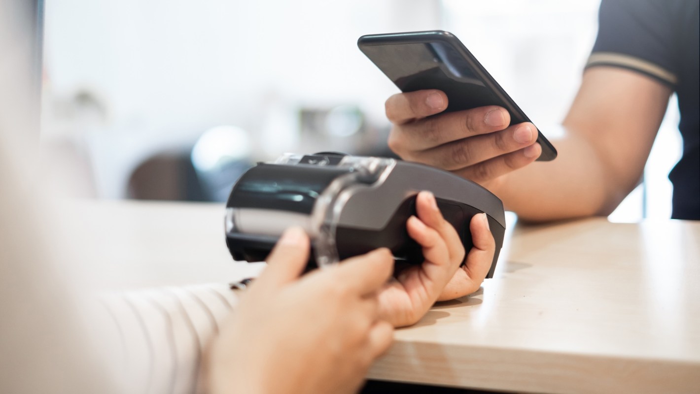 A person holds a mobile phone while another hand uses a payment device on a table.