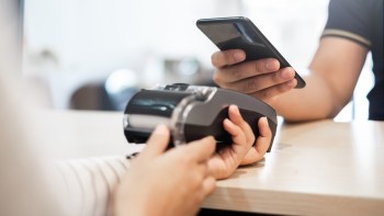 A person holds a mobile phone while another hand uses a payment device on a table.