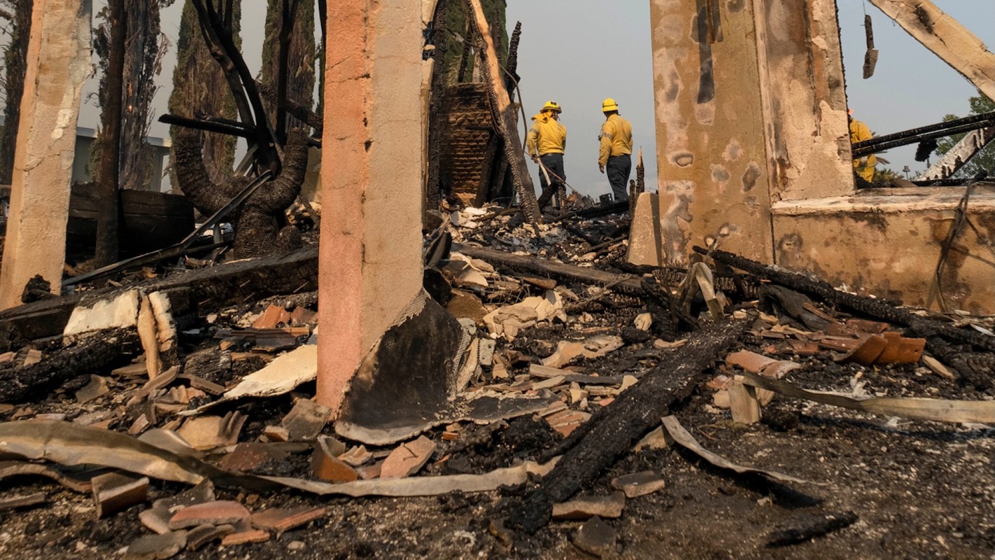 Two firefighters survey the remnants of a building destroyed by fire, surrounded by charred debris and ash.
